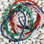 A stack of multi-strand beaded bracelets in various colors including green, blue, red, pink, and white, arranged on a bed of small white pebbles.