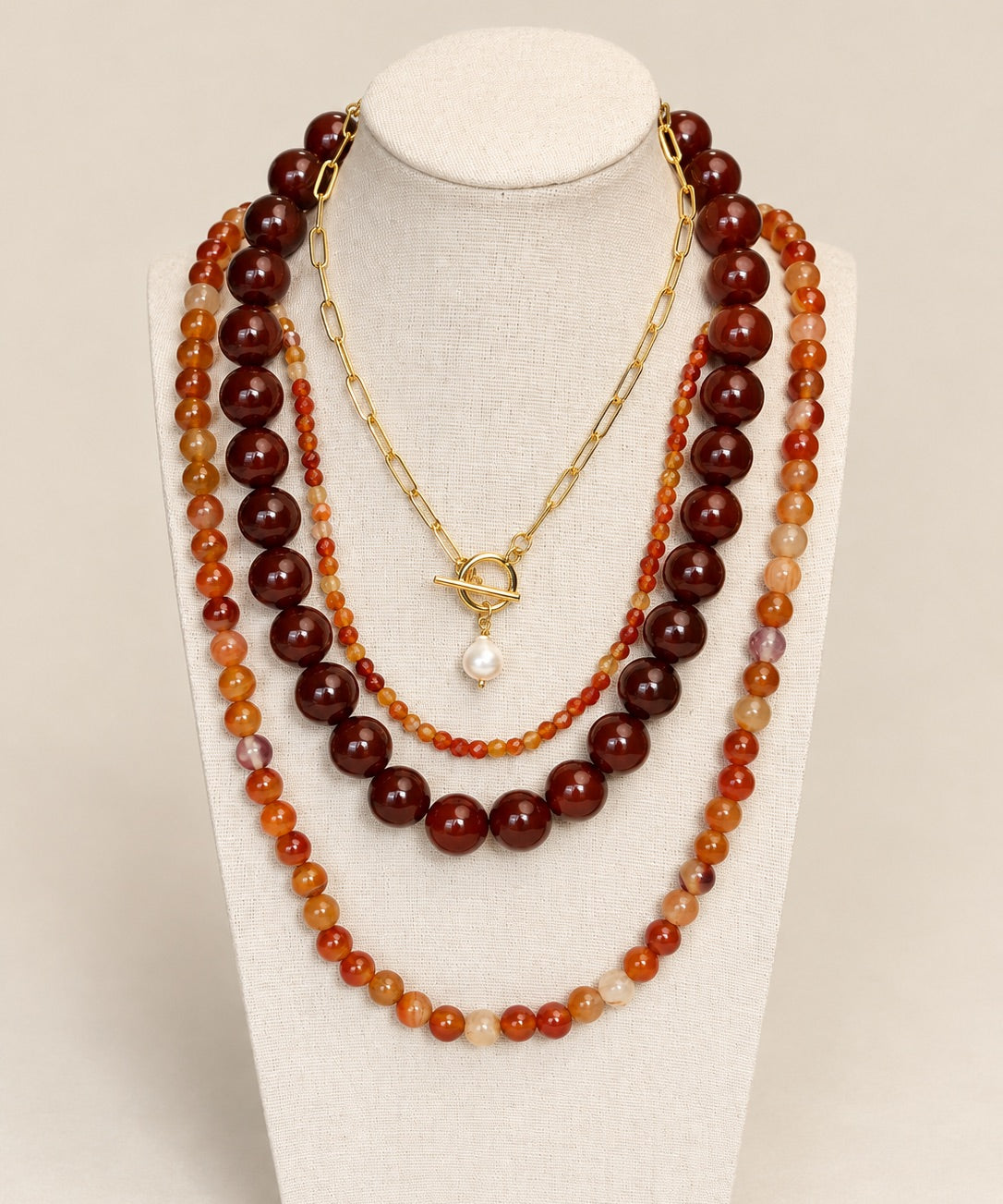A multi-layered necklace set displayed on a cream fabric mannequin bust. The set features three distinct strands: a long strand of dark reddish-brown polished beads, a medium strand of orange and amber marbled beads, and a short gold-tone chain with a toggle clasp and a single white pearl pendant.