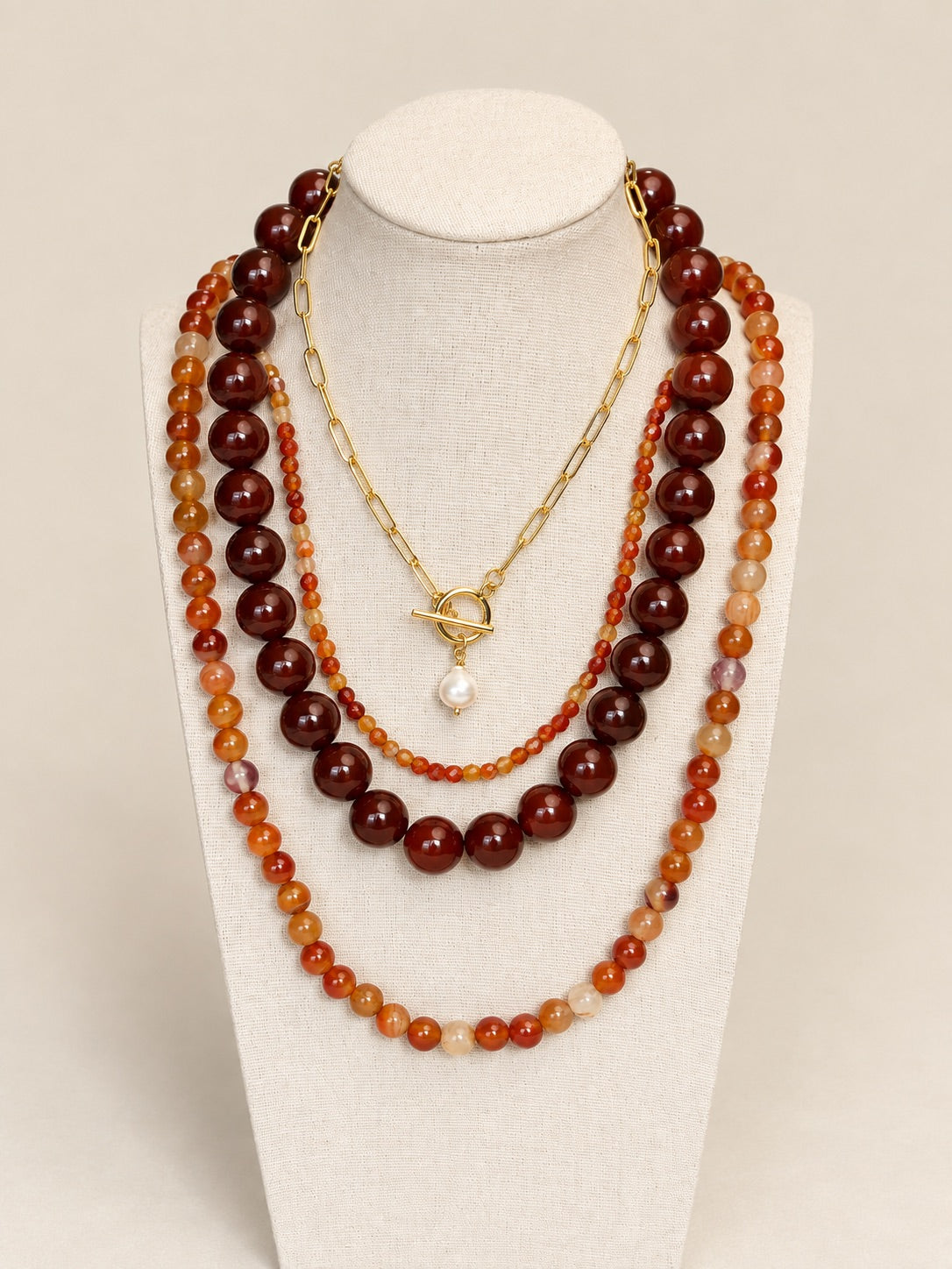 A multi-layered necklace set displayed on a cream fabric mannequin bust. The set features three distinct strands: a long strand of dark reddish-brown polished beads, a medium strand of orange and amber marbled beads, and a short gold-tone chain with a toggle clasp and a single white pearl pendant.