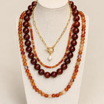 A multi-layered necklace set displayed on a cream fabric mannequin bust. The set features three distinct strands: a long strand of dark reddish-brown polished beads, a medium strand of orange and amber marbled beads, and a short gold-tone chain with a toggle clasp and a single white pearl pendant.