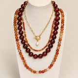 A multi-layered necklace set displayed on a cream fabric mannequin bust. The set features three distinct strands: a long strand of dark reddish-brown polished beads, a medium strand of orange and amber marbled beads, and a short gold-tone chain with a toggle clasp and a single white pearl pendant.
