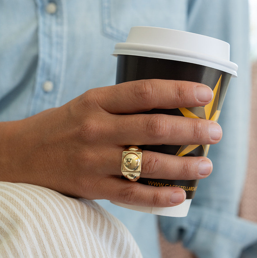 Hand holding a coffee cup with a gold ring, wearing a light blue denim shirt.